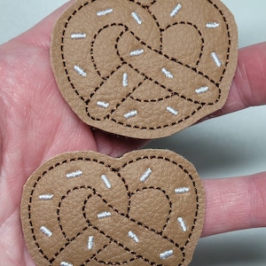 May include: Two brown leather patches with embroidered pretzel shapes. The pretzels are outlined in brown thread and have white sprinkles embroidered on them.