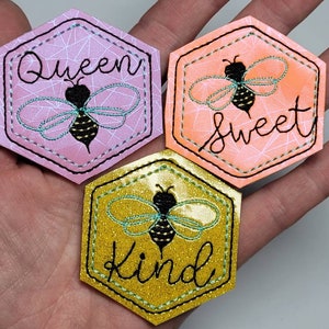 May include: Three hexagon-shaped embroidered patches with a bee design and the words "Queen", "Sweet", and "Kind". The patches are on a pink, orange, and gold background.