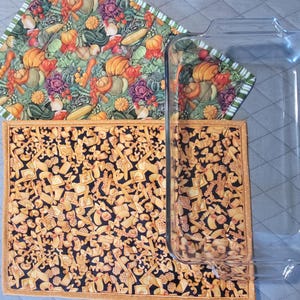 May include: A clear glass baking dish rests on two quilted pot holders. One pot holder features a colorful vegetable print with pumpkins, squash, and carrots. The other has a black background with a pasta pattern.