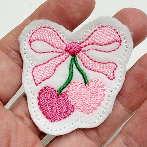 May include: A white felt patch with a pink embroidered bow and two pink cherries. The cherries have green stems.