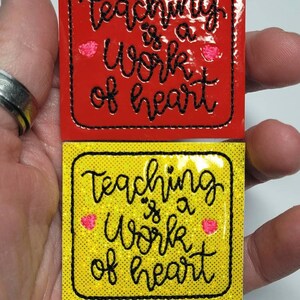 May include: Two iron-on patches with the text "Teaching is a work of heart" in black cursive lettering. The patches are on a red and yellow background with pink hearts.