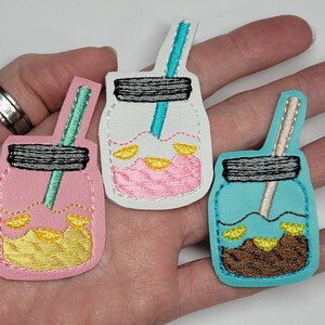 May include: Three embroidered felt mason jar patches with straws and lemon slices. The jars are pink, white, and blue.
