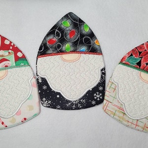 May include: Three Christmas gnome-themed pot holders. The first pot holder has a red background with a white and green pattern of mugs with hot chocolate and marshmallows. The second pot holder has a black background with a white and silver pattern of Christmas lights. The third pot holder has a black background with a red and green pattern of poinsettias.