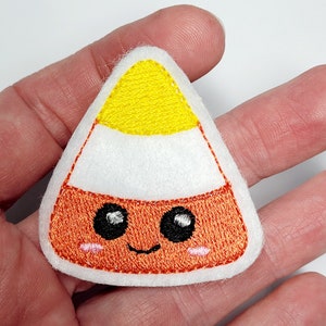 May include: A white felt applique of a cartoon candy corn with orange and yellow felt details. The candy corn has a smiling face with black eyes and pink cheeks.
