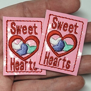 May include: Two pink iron-on patches with red embroidered text that reads "Sweet Hearts" and a red embroidered heart with a blue and green design inside.