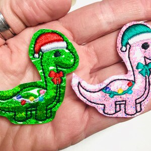 May include: Two sparkly Christmas dinosaur ornaments. One is green with a red Santa hat and the other is pink with a green Santa hat. Both have a green holly wreath around their necks.