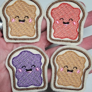 May include: Four embroidered felt patches shaped like slices of bread with different colored fillings. The fillings are brown, red, purple, and brown. Each slice has a smiling face with pink cheeks.