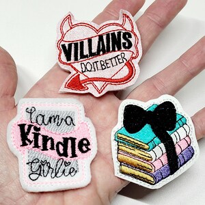 May include: Three embroidered iron-on patches. One patch is a red heart with devil horns and a tail, with the text "VILLAINS DO IT BETTER". Another patch is white with gray and pink accents and the text "I'm a Kindle Girl". The third patch is white with a stack of colorful books tied with a black bow.