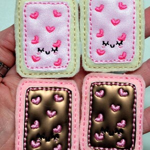 May include: Four felt patches, two pink and two brown, with a white border and pink hearts. The pink patches have a pink heart pattern and a pink smiling face with closed eyes. The brown patches have a brown heart pattern and a pink smiling face with closed eyes.