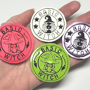 May include: Four round patches with the text "BASIC WITCH" and a witch illustration. The patches are in pink, white, green, and purple. The witch is wearing a hat and has long hair. The patches are surrounded by a black border.