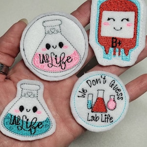 May include: Four embroidered iron-on patches with a science theme. One patch features a beaker with the text "Lab Life" in pink. Another patch features a beaker with the text "Lab Life" in blue. A third patch features a blood bag with the text "B+" and a smiling face. The final patch features three beakers and the text "We Don't Guess Lab Life".