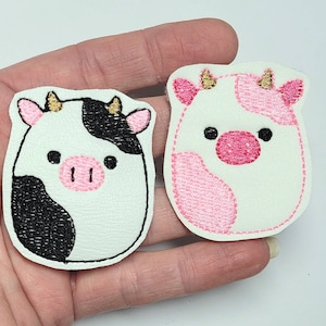 May include: Two felt cow appliques, one black and white and one pink and white. Both have gold glitter horns and pink noses.