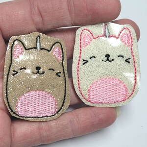 May include: Two glittery cat-shaped patches. One is tan with pink accents, the other is off-white with pink accents. Both have a unicorn horn and are outlined in black. The patches are smiling.