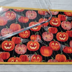 May include: A black quilted hot pad with a yellow border features a repeating pattern of orange jack-o'-lantern faces and spiderwebs. A clear glass baking dish is on top of the hot pad.