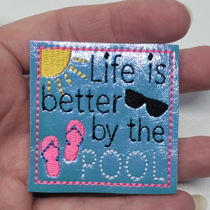 May include: Square, turquoise patch with the embroidered phrase "Life is better by the pool." The patch features a yellow sun, black sunglasses, and pink flip-flops. The design is outlined with a pink border.