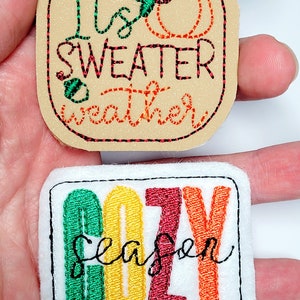 May include: Two embroidered patches with fall themes. One is tan with the text "It's sweater weather" and a pumpkin design. The other is white with the word "COZY season" in colorful letters.