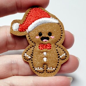 May include: A gingerbread man with a red and white Santa hat, wearing a red bow tie and white buttons. The gingerbread man is embroidered on a brown felt-like fabric.