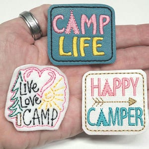 May include: Three embroidered iron-on patches with a camping theme. The first patch is blue with pink and yellow embroidery and says "Camp Life". The second patch is white with pink, yellow, green, and black embroidery and says "Live Love Camp". The third patch is white with pink, gold, and blue embroidery and says "Happy Camper".