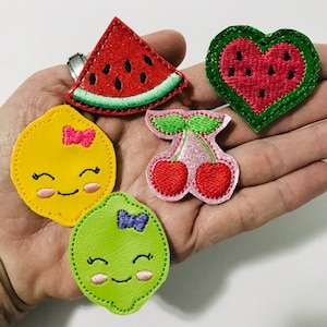 May include: Five felt fruit patches, including a watermelon slice, a heart-shaped watermelon, a lemon with a pink bow, a lime with a purple bow, and two cherries with green stems and pink leaves.