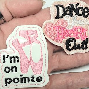 May include: Two iron-on patches for clothing. One patch is pink and white and says "I'm on pointe" with a pink ballet shoe. The other patch is pink and white and says "Dance your heart out".