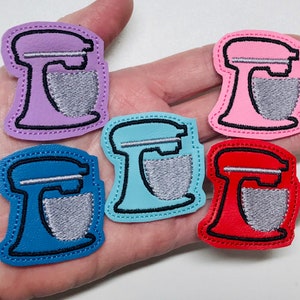 May include: Five felt appliques of a stand mixer in different colors: purple, blue, pink, red, and teal. Each applique has a black outline and a silver-colored mixing bowl.