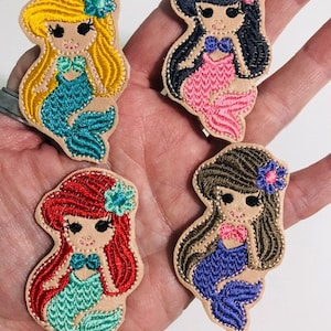 May include: Four embroidered iron-on patches featuring mermaid characters with different hair colors and tail colors. The mermaids are wearing seashell bras and have flowers in their hair.