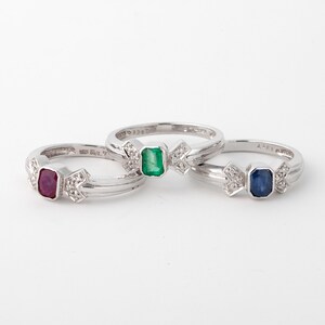 Op de afbeelding: Drie zilveren ringen met een strikdesign en een verschillende gekleurde edelsteen in elke ring. De ringen hebben een robijn, een smaragd en een saffier.