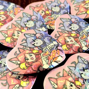 Op de afbeelding: Een verzameling hartvormige buttons met een kleurrijke anime-illustratie van twee personages met kattenoren. Het ene personage heeft rood haar en het andere blauw haar. De tekst "MAD MEW" staat op elke button gedrukt.