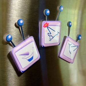May include: Three cartoon-style magnets with a retro TV design. Each magnet has a light purple frame, white screen, and a blue antenna with a blue ball on top. The screens feature different cartoon faces and designs.