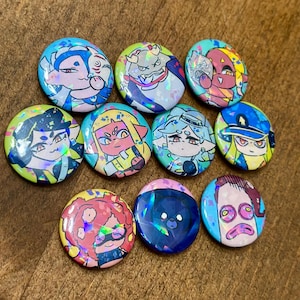 K&ouml;nnte beinhalten: Eine Sammlung von zehn bunten, runden Ansteckbuttons mit holografischen Akzenten. Jeder Button zeigt ein einzigartiges Cartoon-Charakter-Design mit leuchtenden Farben wie Blau, Gelb, Rosa und Gr&uuml;n. Die Buttons sind auf einer Holzoberfl&auml;che angeordnet.