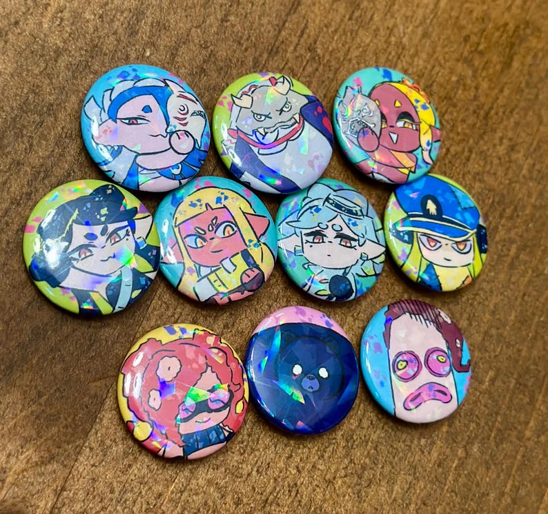 K&ouml;nnte beinhalten: Eine Sammlung von zehn bunten, runden Ansteckbuttons mit holografischen Akzenten. Jeder Button zeigt ein einzigartiges Cartoon-Charakter-Design mit leuchtenden Farben wie Blau, Gelb, Rosa und Gr&uuml;n. Die Buttons sind auf einer Holzoberfl&auml;che angeordnet.