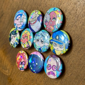 K&ouml;nnte beinhalten: Elf ovale Kn&ouml;pfe mit Illustrationen von Cartoon-Charakteren in verschiedenen Farben. Die Kn&ouml;pfe zeigen Charaktere in Blau, Rosa, Gelb und Gr&uuml;n mit holografischen Details. Die Kn&ouml;pfe sind auf einer braunen Oberfl&auml;che angeordnet.