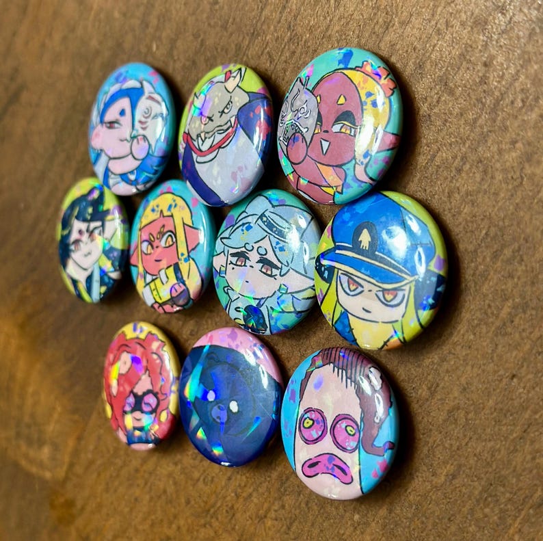 K&ouml;nnte beinhalten: Elf ovale Kn&ouml;pfe mit Illustrationen von Cartoon-Charakteren in verschiedenen Farben. Die Kn&ouml;pfe zeigen Charaktere in Blau, Rosa, Gelb und Gr&uuml;n mit holografischen Details. Die Kn&ouml;pfe sind auf einer braunen Oberfl&auml;che angeordnet.