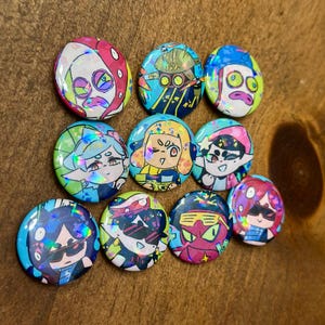 K&ouml;nnte beinhalten: Eine Sammlung von zehn runden, farbenfrohen Kn&ouml;pfen mit holografischen Designs von Cartoon-Charakteren. Die Kn&ouml;pfe haben eine gl&auml;nzende Oberfl&auml;che und eine Vielzahl von Farben, darunter Rosa, Blau, Gr&uuml;n und Gelb. Die Charaktere tragen Sonnenbrillen.