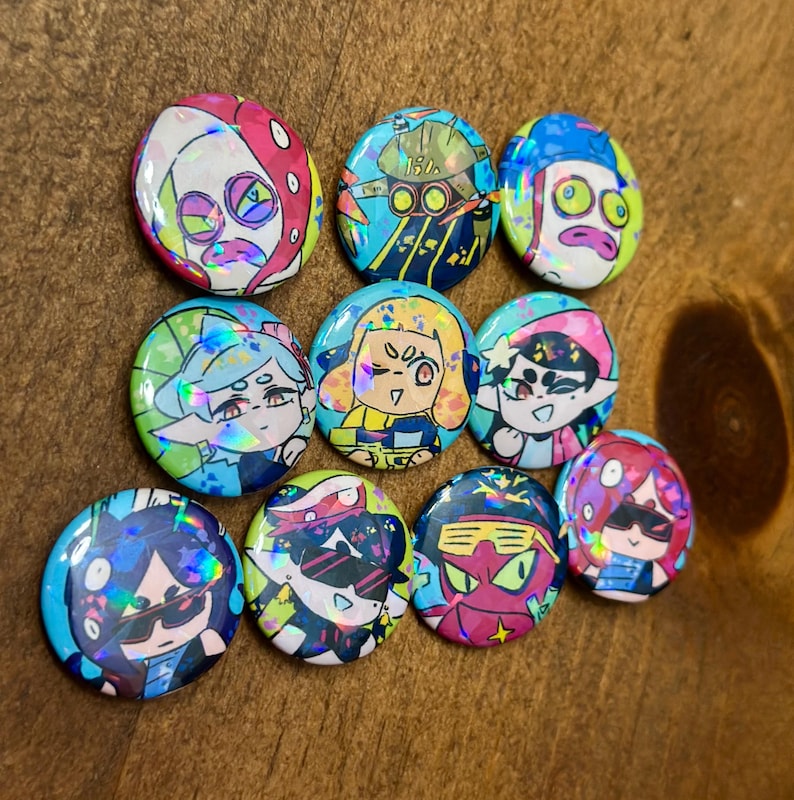 K&ouml;nnte beinhalten: Eine Sammlung von zehn runden, farbenfrohen Kn&ouml;pfen mit holografischen Designs von Cartoon-Charakteren. Die Kn&ouml;pfe haben eine gl&auml;nzende Oberfl&auml;che und eine Vielzahl von Farben, darunter Rosa, Blau, Gr&uuml;n und Gelb. Die Charaktere tragen Sonnenbrillen.