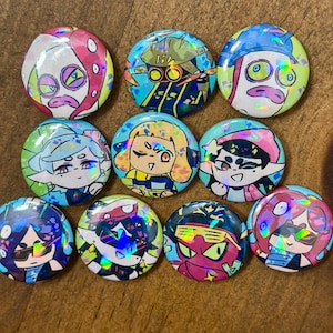 K&ouml;nnte beinhalten: Eine Sammlung von zehn bunten, runden Kn&ouml;pfen mit holografischen Cartoon-Charakter-Designs. Die Kn&ouml;pfe zeigen verschiedene Charaktere in leuchtenden Farben, darunter Pink, Blau, Gelb und Gr&uuml;n, auf einer Holzoberfl&auml;che.