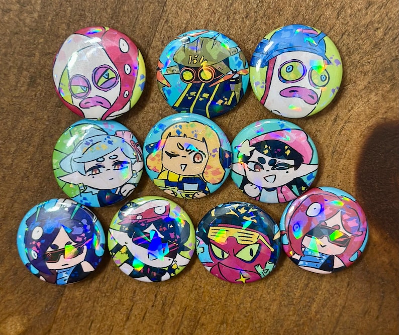 K&ouml;nnte beinhalten: Eine Sammlung von zehn bunten, runden Kn&ouml;pfen mit holografischen Cartoon-Charakter-Designs. Die Kn&ouml;pfe zeigen verschiedene Charaktere in leuchtenden Farben, darunter Pink, Blau, Gelb und Gr&uuml;n, auf einer Holzoberfl&auml;che.