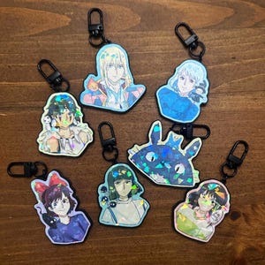 Puede incluir: Una colección de llaveros holográficos de personajes de anime con cierres negros. Los llaveros presentan varios personajes en colores vibrantes, incluyendo una criatura azul con forma de gato y figuras con rasgos faciales y ropa detallados.