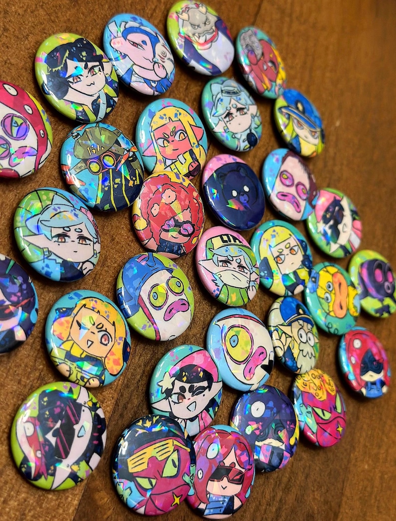 K&ouml;nnte beinhalten: Eine Sammlung bunter, runder Ansteckbuttons mit holografischen Akzenten. Jeder Button zeigt ein einzigartiges Cartoon-Charakter-Design mit leuchtenden Farben und detaillierten Kunstwerken. Die Buttons sind auf einer Holzoberfl&auml;che angeordnet.