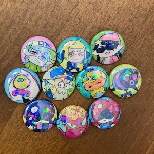 K&ouml;nnte beinhalten: Eine Sammlung von zehn runden Ansteckbuttons mit Cartoon-Charakteren und holografischen Effekten. Jeder Button hat ein einzigartiges Design mit leuchtenden Farben wie Rosa, Blau, Gr&uuml;n und Gelb. Auf einer Holzoberfl&auml;che angeordnet.