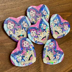 May include: Five heart-shaped pin-back buttons featuring a holographic finish. Each button displays a colorful illustration of three animated characters: a person with long red hair, a person with blue hair, and a tan cat-like creature with a gold coin.