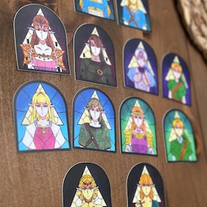 Puede incluir: Un conjunto de 11 pegatinas ilustradas con diferentes personajes de la serie de videojuegos The Legend of Zelda. Las pegatinas son de estilo vitral con un diseño triangular y presentan personajes como Link, Zelda y Ganondorf.