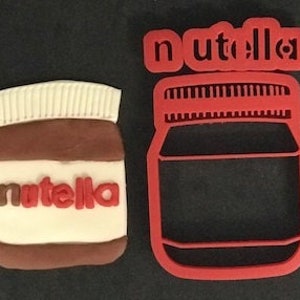 May include: Red plastic cookie cutter shaped like a jar of Nutella with the word "Nutella" printed on the jar. The cookie cutter is next to a brown and white cookie decorated with the word "Nutella".