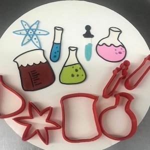 May include: A collection of science-themed cookie cutters and decorated cookies. The cookies feature colorful designs of beakers, test tubes, and an atom symbol. The cookie cutters are red plastic.