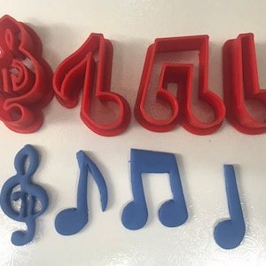 May include: Red plastic cookie cutters in the shapes of a treble clef and musical notes are displayed above blue fondant versions of the same shapes. The cutters are for baking and decorating cookies with a music theme.