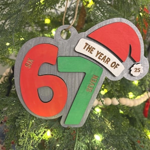 May include: Wooden Christmas ornament with the text "THE YEAR OF" and the numbers "67" in red and green. A red Santa hat and the number "25" are also featured. The ornament is hanging on a Christmas tree.