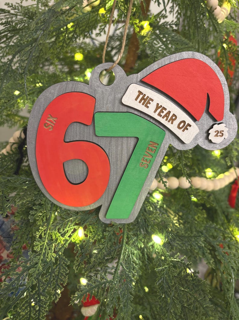 May include: Wooden Christmas ornament with the text "THE YEAR OF" and the numbers "67" in red and green. A red Santa hat and the number "25" are also featured. The ornament is hanging on a Christmas tree.