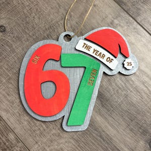 May include: Wooden Christmas ornament with the text "THE YEAR OF 67" and a Santa hat. The numbers are red and green, with the word "SIX" on the number 6 and "SEVEN" on the number 7. The ornament has a silver-colored border and a twine loop for hanging.