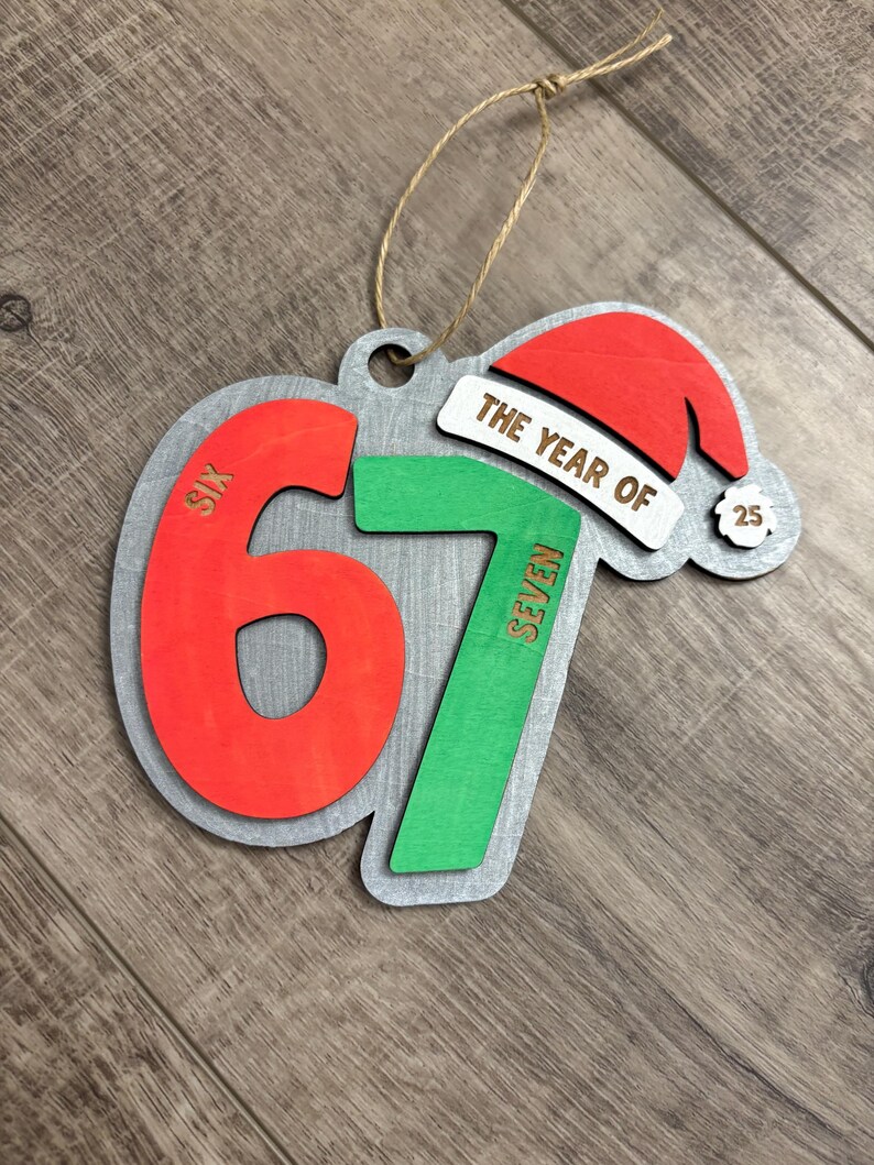May include: Wooden Christmas ornament with the text "THE YEAR OF 67" and a Santa hat. The numbers are red and green, with the word "SIX" on the number 6 and "SEVEN" on the number 7. The ornament has a silver-colored border and a twine loop for hanging.