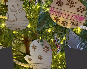 Puede incluir: Adornos navideños de madera con forma de muñeco de nieve, un guante y una bola con el nombre "Kennedy" y el año "2025" grabados. Los adornos cuelgan de un árbol de Navidad con luces y cuentas blancas.